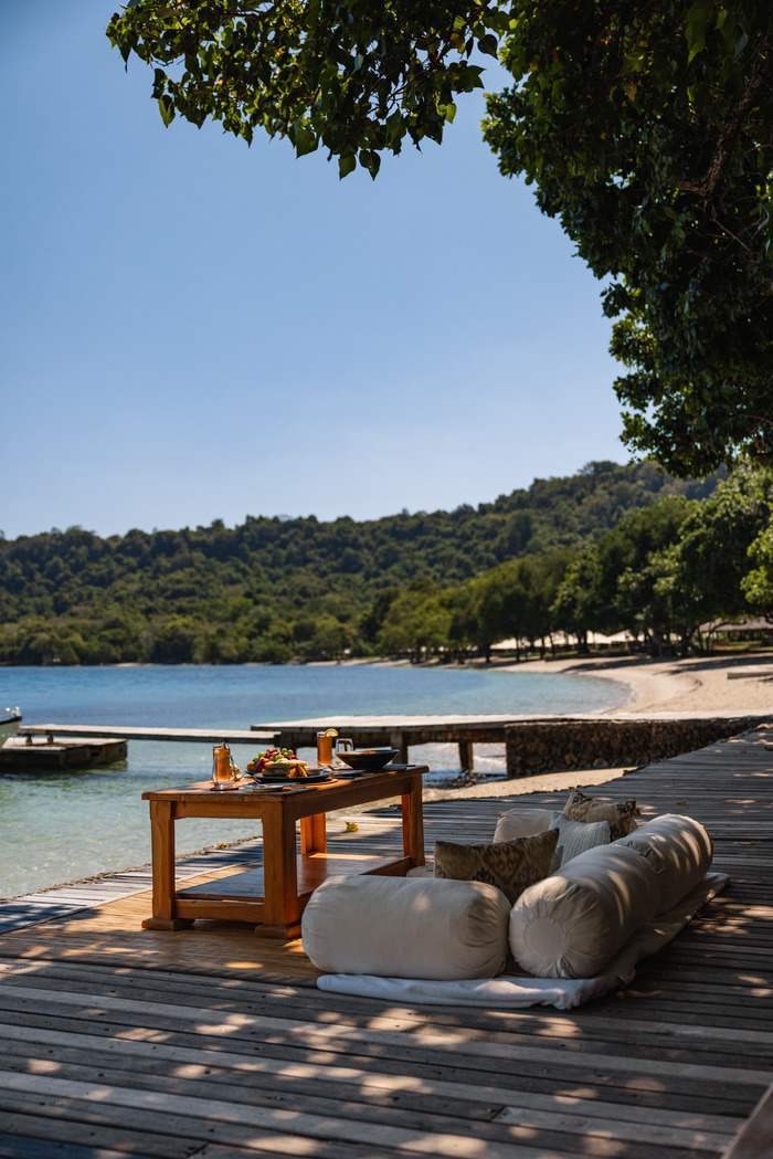 Outdoor dining setup on a wooden deck with floor cushions overlooking a turquoise bay and forested hills.
