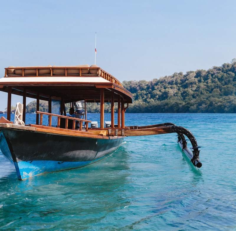 A wooden outrigger boat with a canopy roof on clear blue water near a green island.