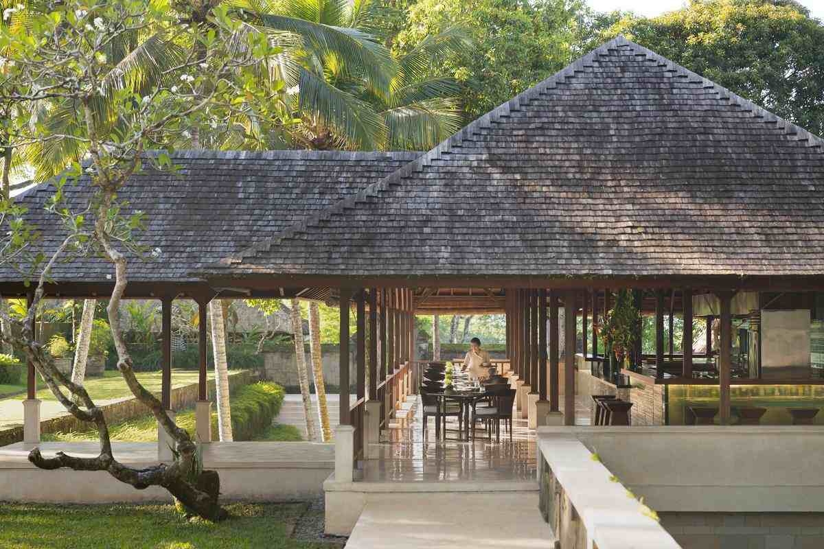 An open-air dining pavilion with set tables and chairs under a massive tiled roof, surrounded by tropical palm trees.