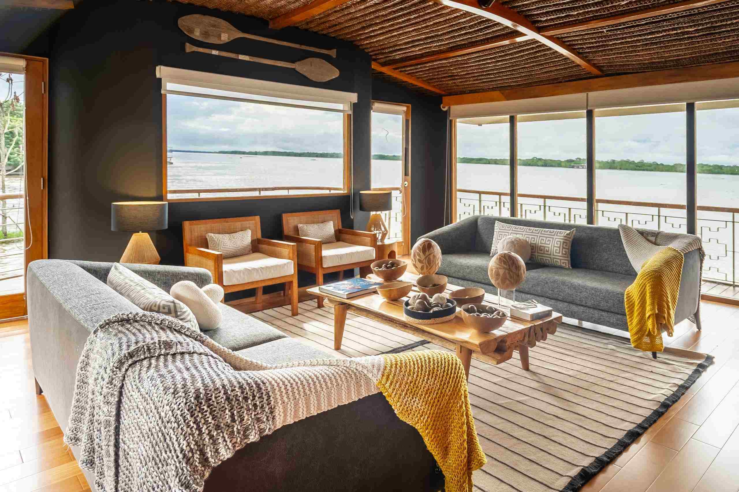 Delfin I's lounge with grey sofas, wooden armchairs, and floor-to-ceiling windows showing a wide river landscape.