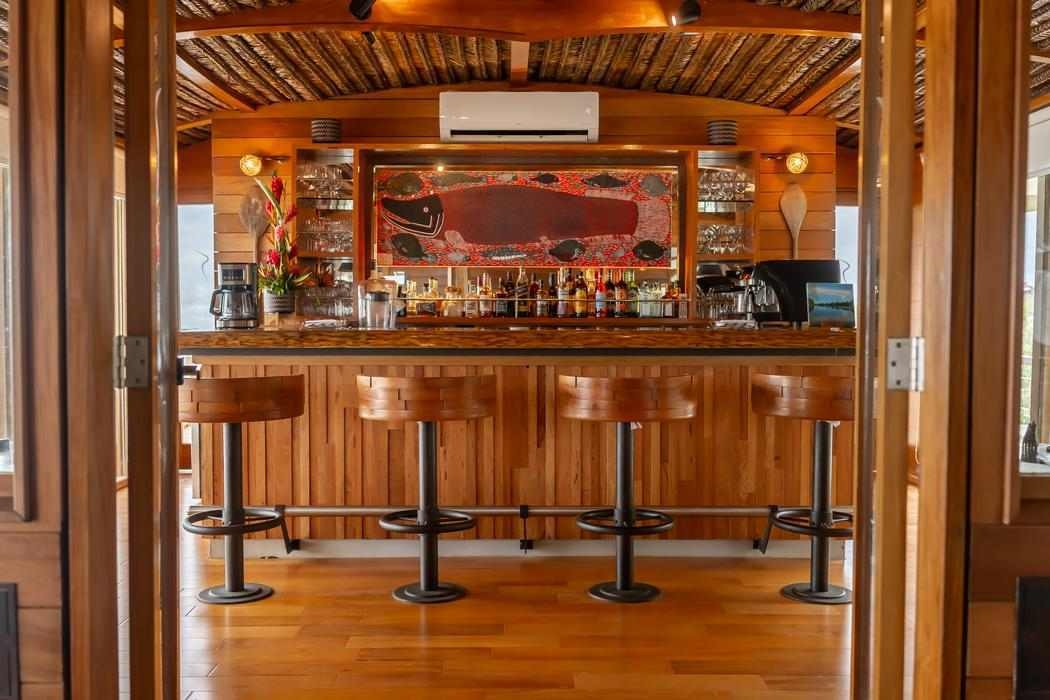 A wooden bar with four stools and shelves of bottles under a thatched ceiling on Delfin I.