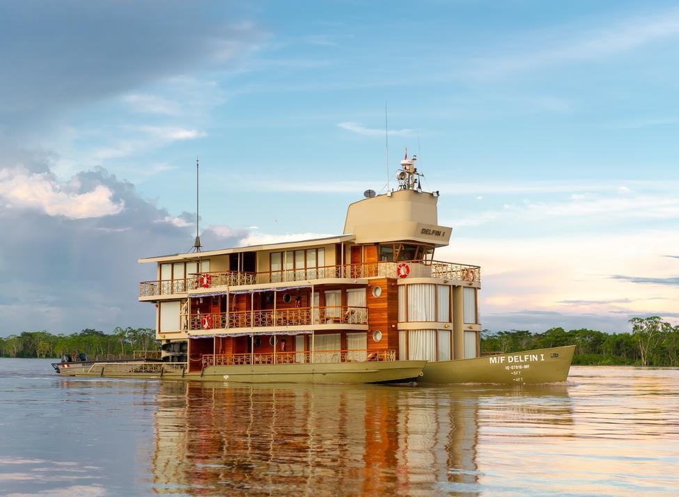 Side view of Delfin I luxury riverboat sailing on calm water with a forest background at sunset.