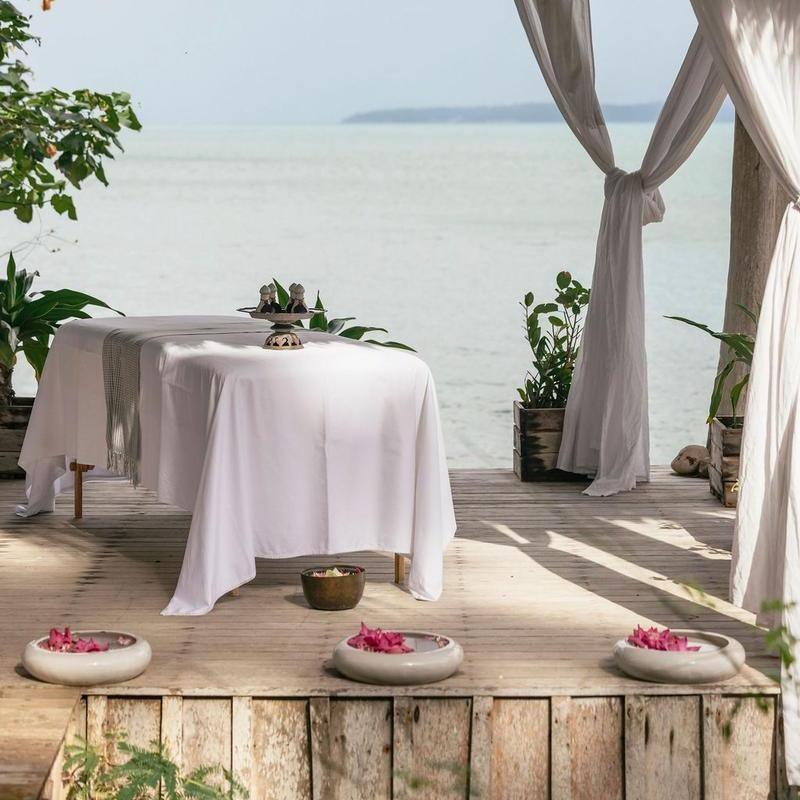 An outdoor massage table on a wooden pier overlooking the ocean with decorative flower bowls.