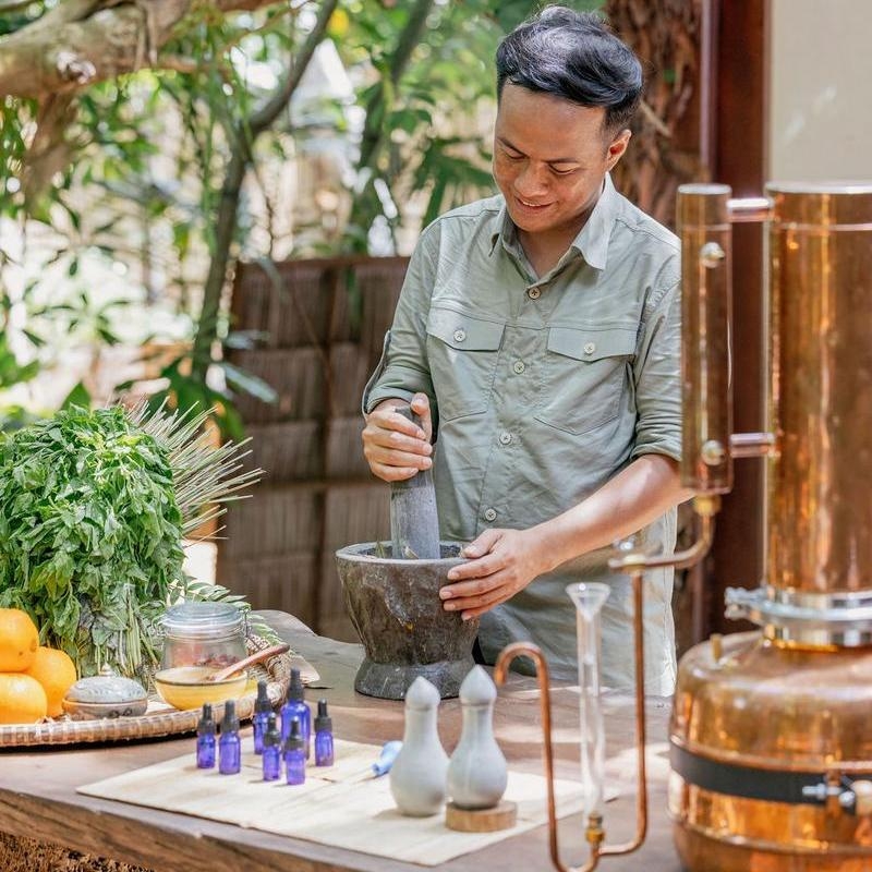 A person grinding herbs in a mortar and pestle next to blue glass bottles and copper spa equipment.