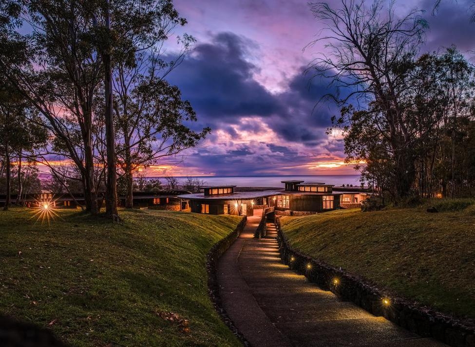 A lighted modern hotel building at dusk with a stone path leading toward it under a vibrant purple and orange sunset sky.