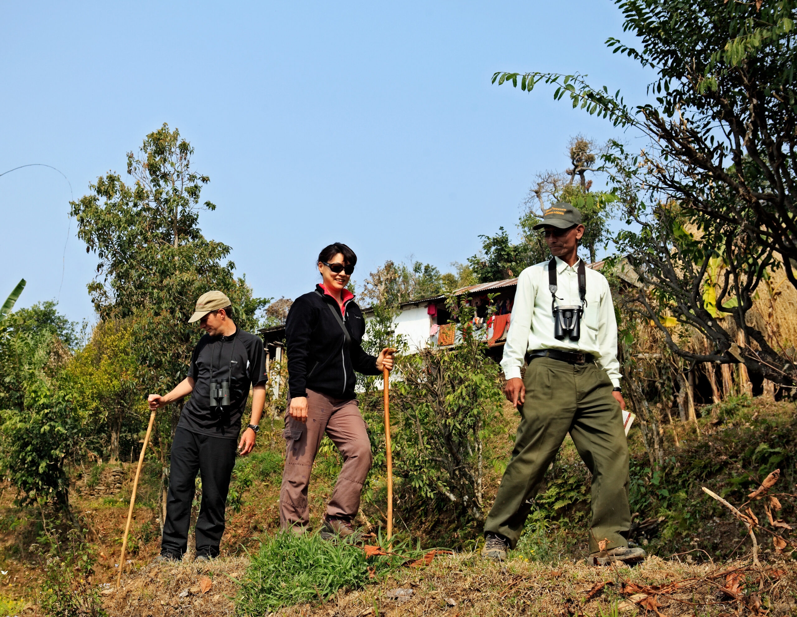 Three individuals with walking sticks standing in a natural landscape with trees and clear skies.