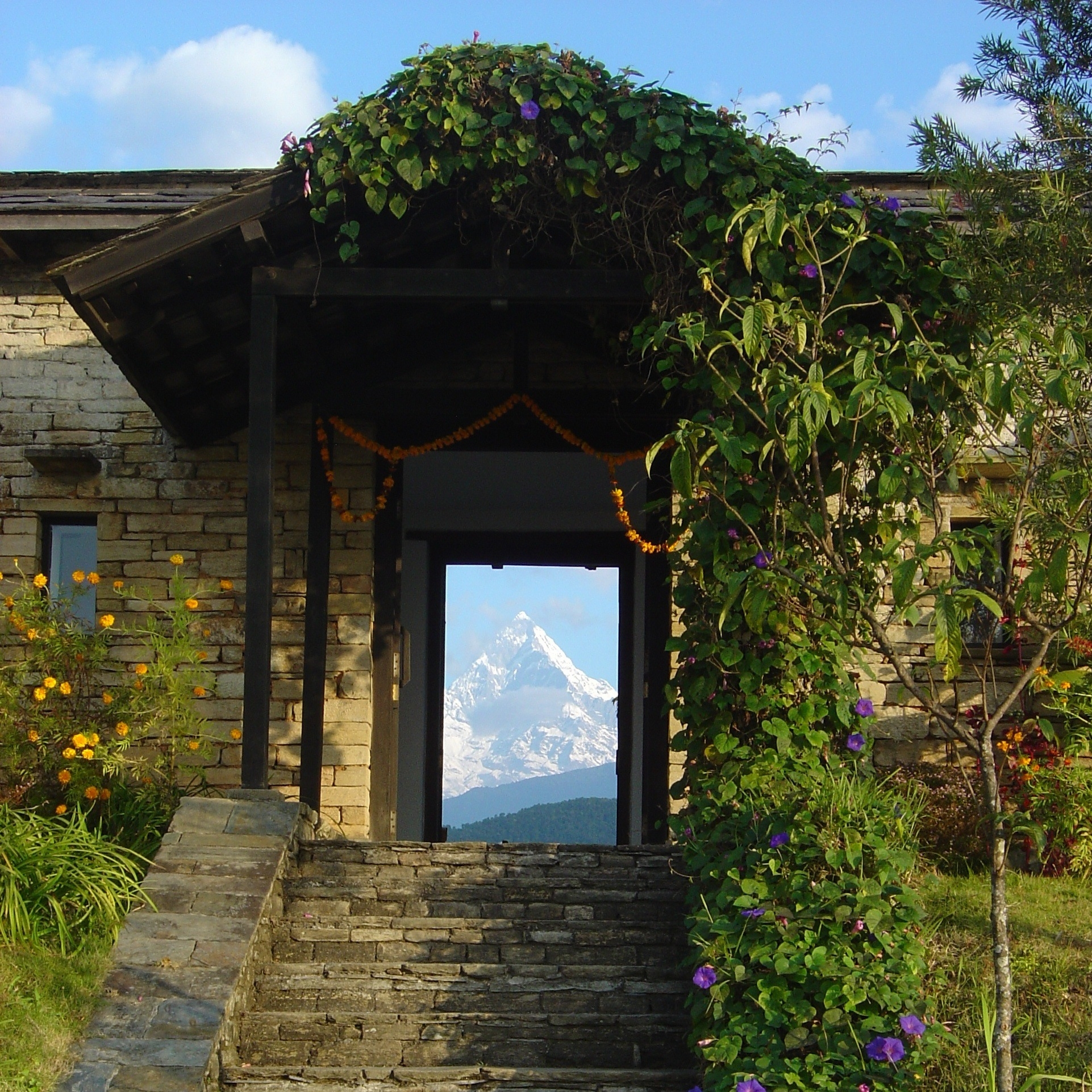 Stone house entrance framing a distant snow-capped mountain peak, adorned with flowers and vines.