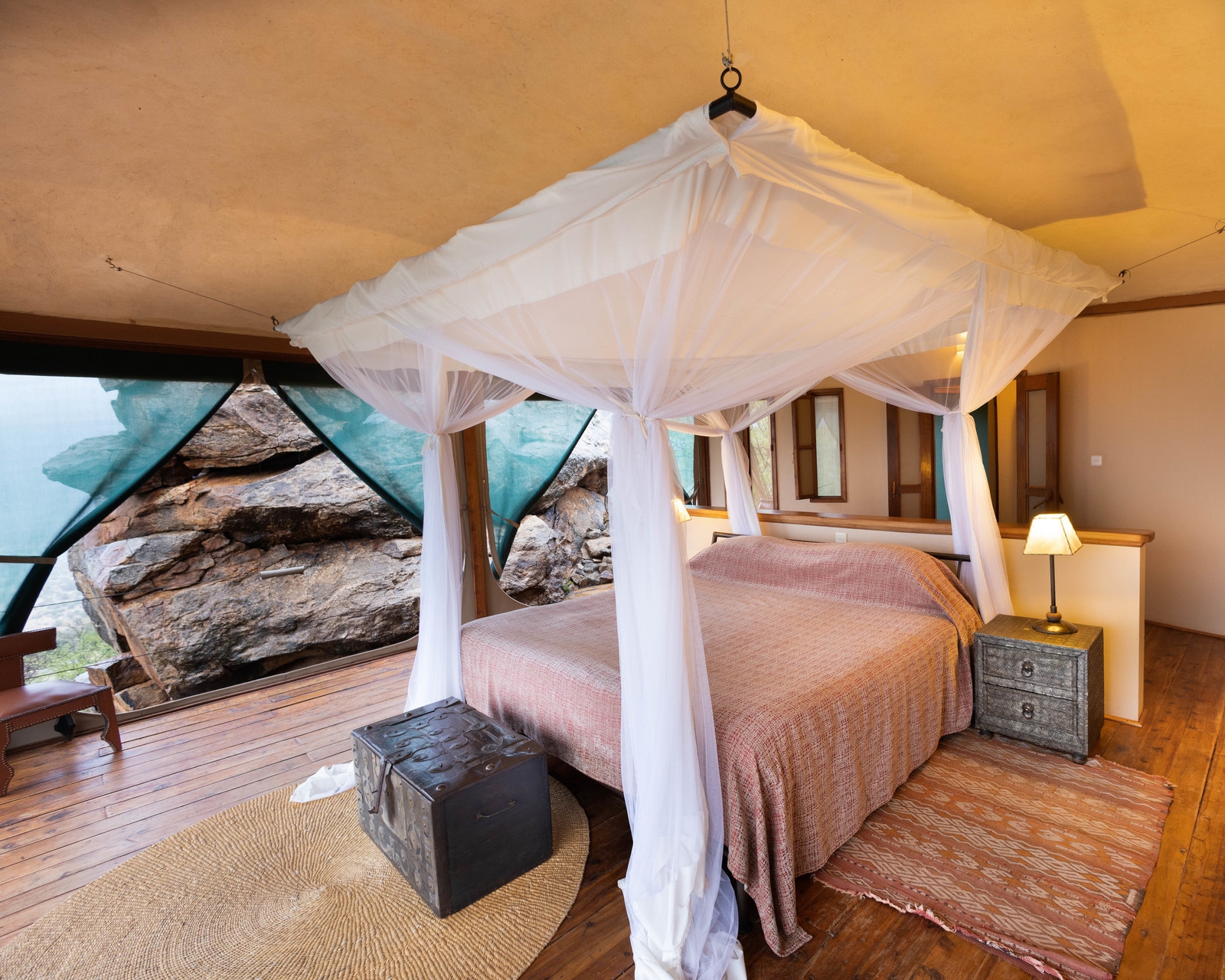 A luxury bedroom with wooden floors, a canopy bed, and a natural boulder wall inside a suite at Saruni Samburu.