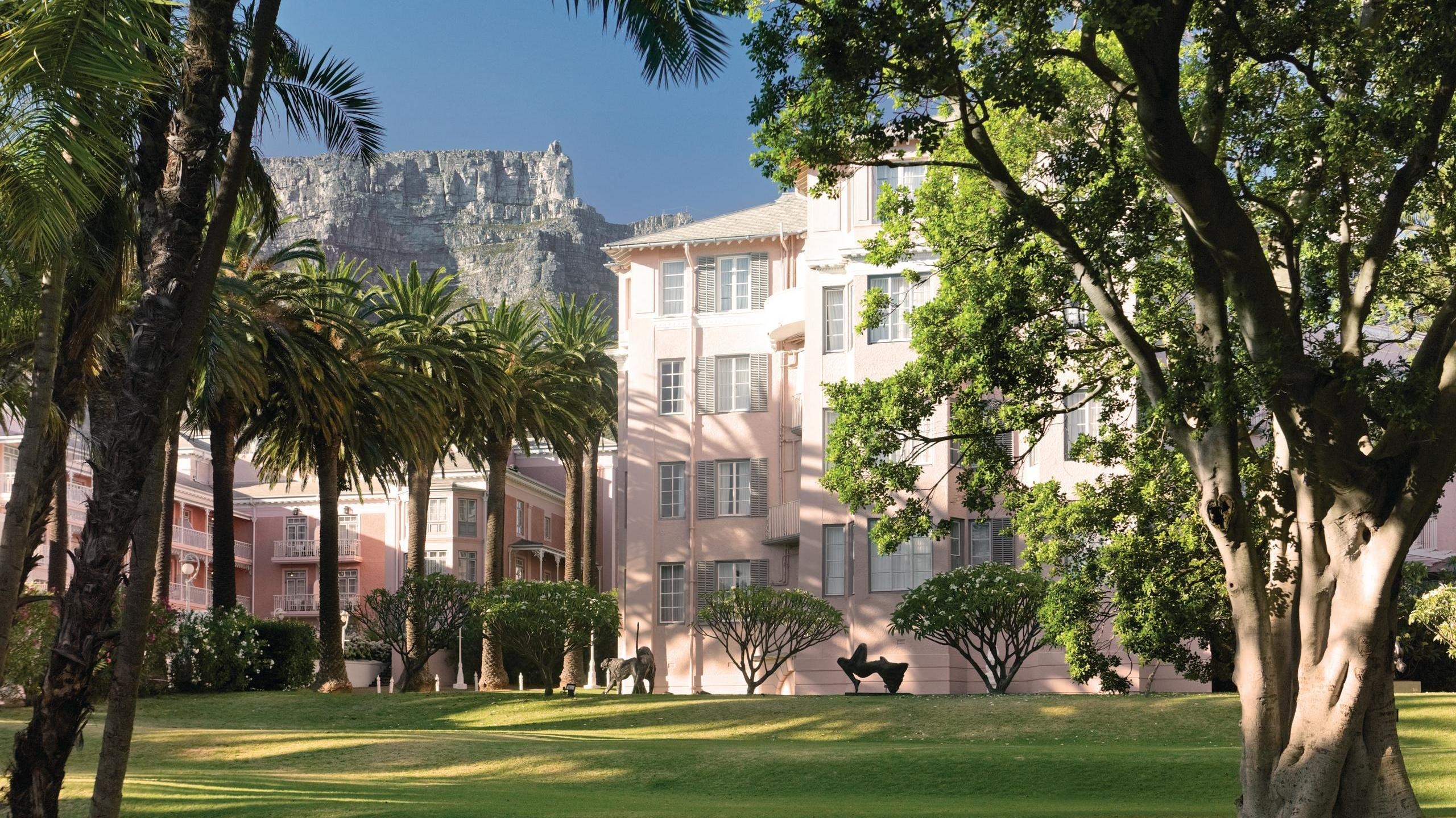 Lush green gardens and palm trees in front of the classic pink Mount Nelson hotel building with mountains behind.
