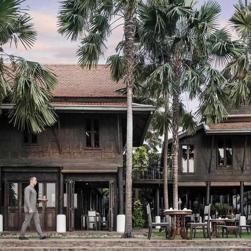 Dark wood traditional Thai architecture buildings set among lush palm trees with outdoor patio dining furniture.