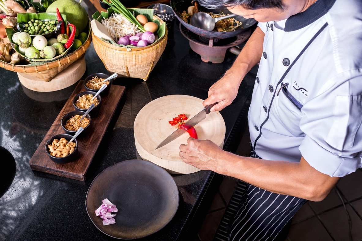 Overhead view of a chef in a white uniform slicing a red chili on a round wooden cutting board.