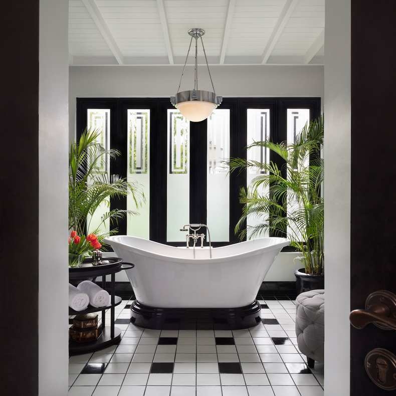 A white clawfoot bathtub in a monochromatic bathroom with checkered tiles, large windows, and indoor palm plants.