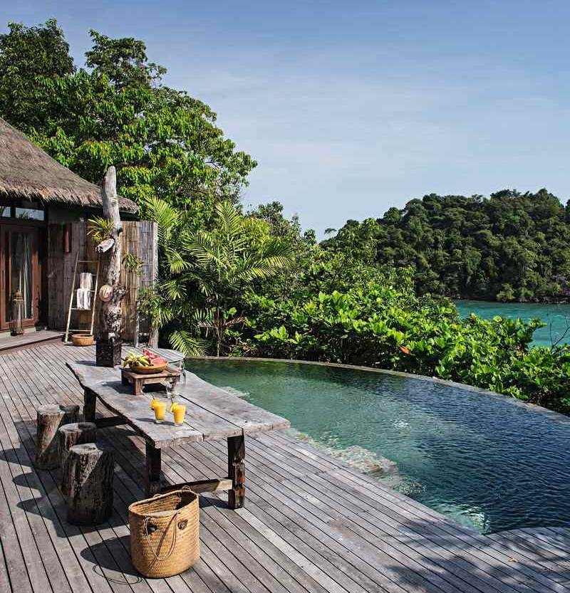 A wooden deck with a swimming pool and dining area surrounded by tropical trees and ocean views.