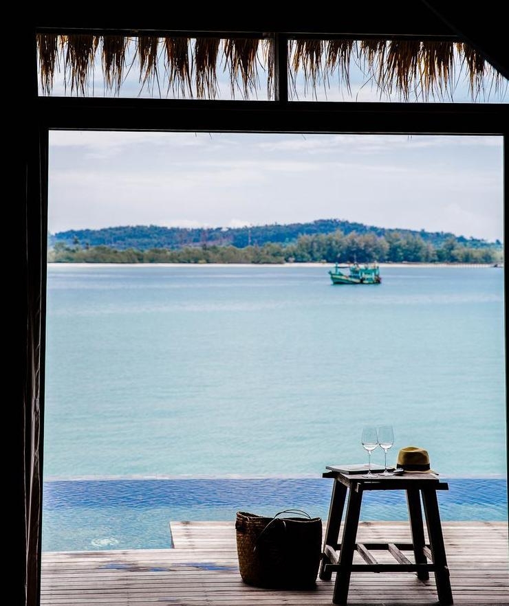 View from a villa deck with an infinity pool, a small table with glasses and a boat on the blue sea.