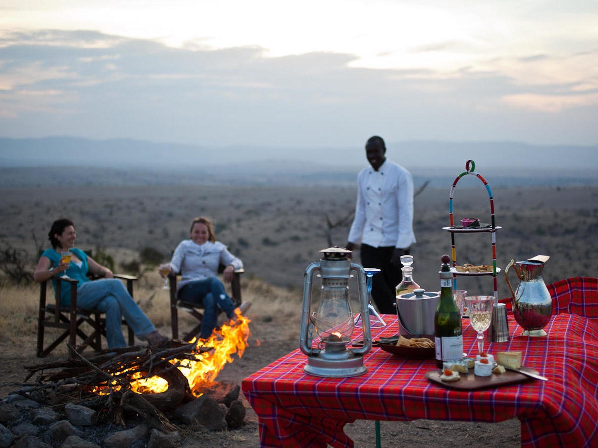 People sitting by a campfire at Lewa House with a table of drinks and snacks overlooking the savanna.