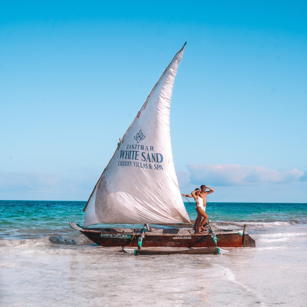 A traditional sailboat on a beach with a person standing on it, under a clear blue sky.