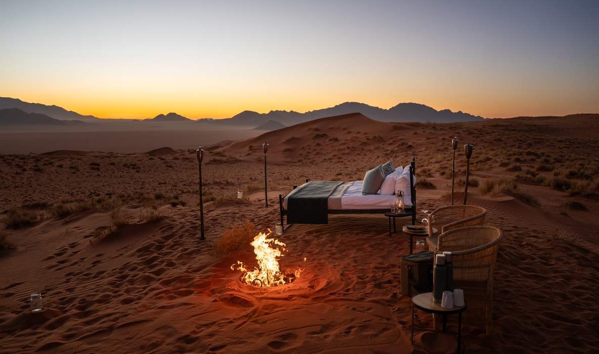 A bed and campfire situated on a red sand dune overlooking a desert valley at sunset.
