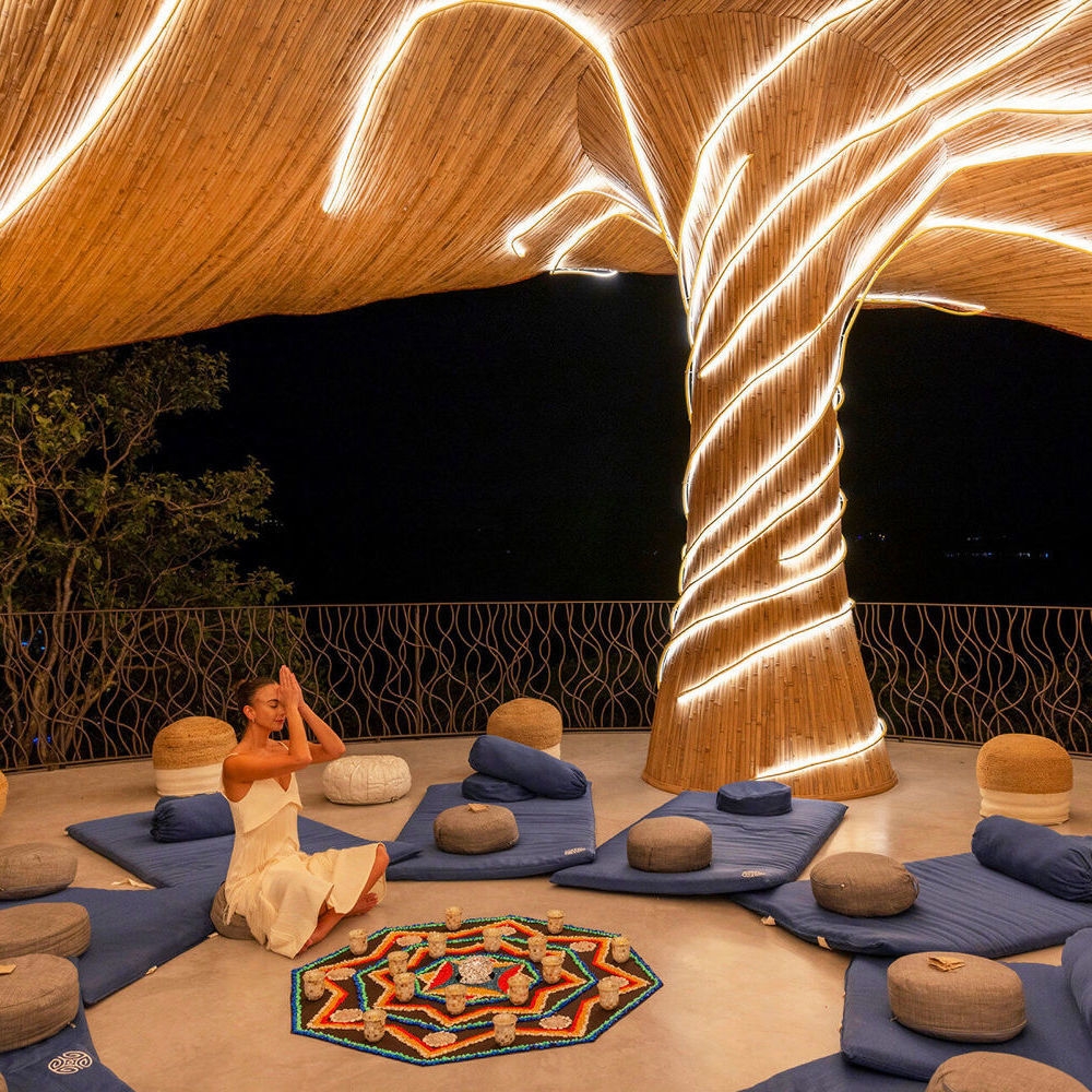 A woman in white sitting on a blue mat meditating inside a sculptural bamboo pavilion with glowing lights.