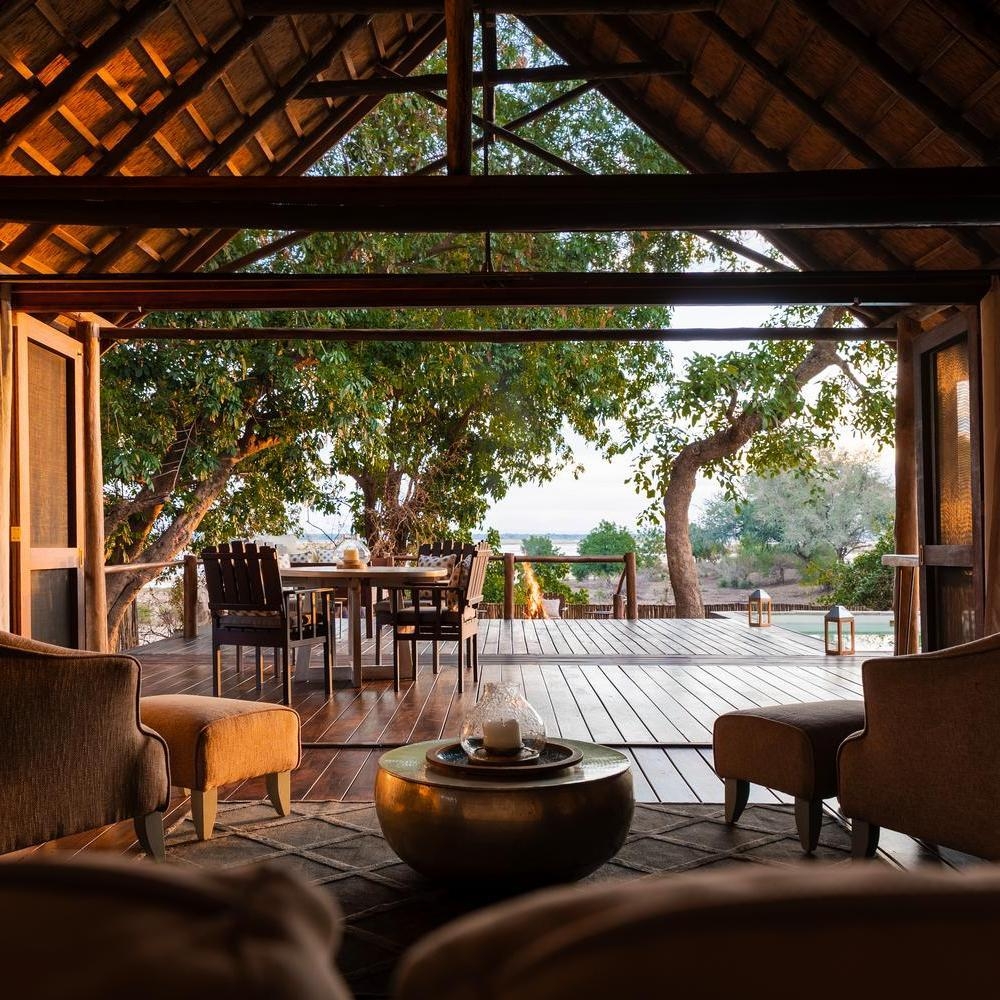 Safari suite lounge opening to a wooden deck with a dining table and fire pit overlooking the river at Chiawa Camp.