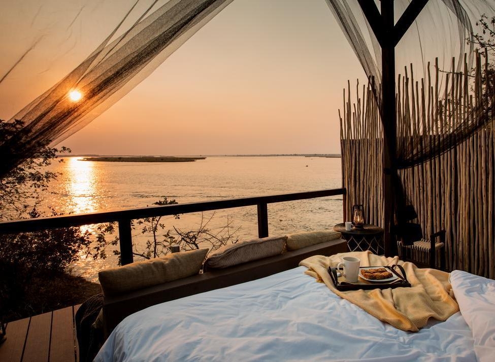View from an outdoor bed with mosquito netting and a breakfast tray, looking over a wide river at sunrise.