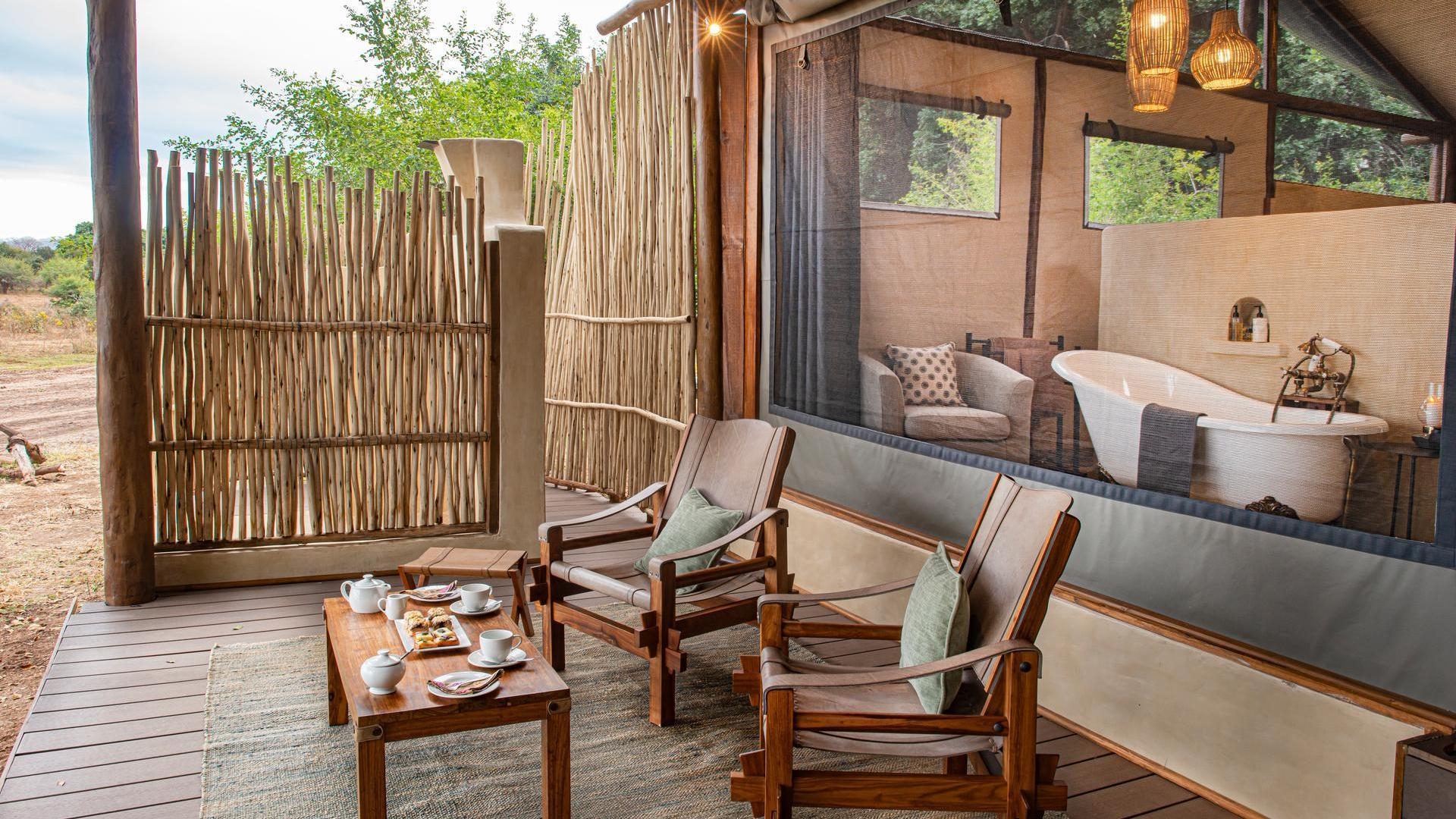 Covered wooden veranda with tea service and chairs, looking through a screen into a bathroom with a freestanding tub.