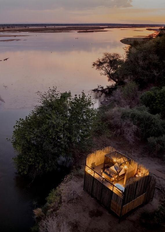 Aerial view of a glowing, open-air wooden tower with a bed inside, sitting on a riverbank at sunset.