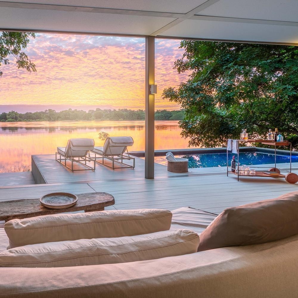 View from a lounge to a wooden deck with a plunge pool, chairs, and drink cart overlooking a river at sunset.