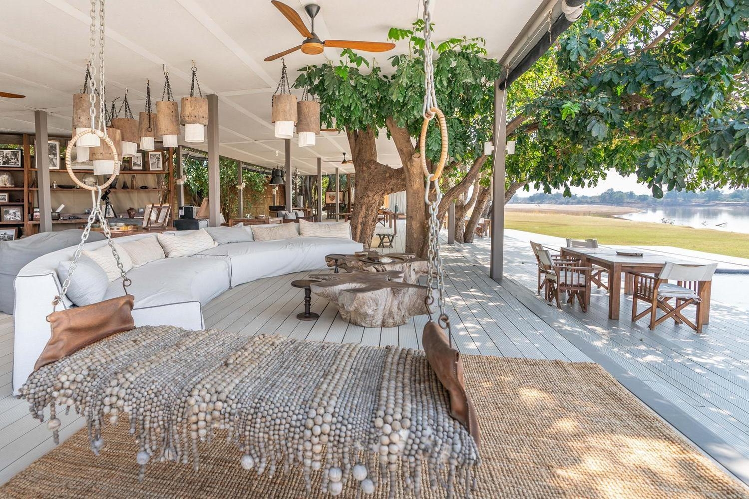 Open-air lounge with a large curved white sofa, rustic wooden tables, and a hanging daybed overlooking a river.