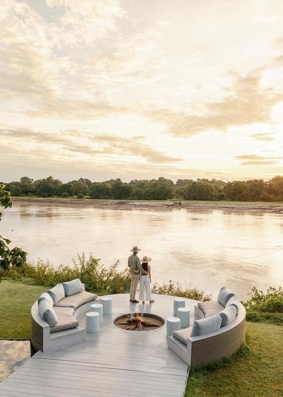 Couple standing by a circular fire pit with curved seating, looking out over a wide river at sunset.