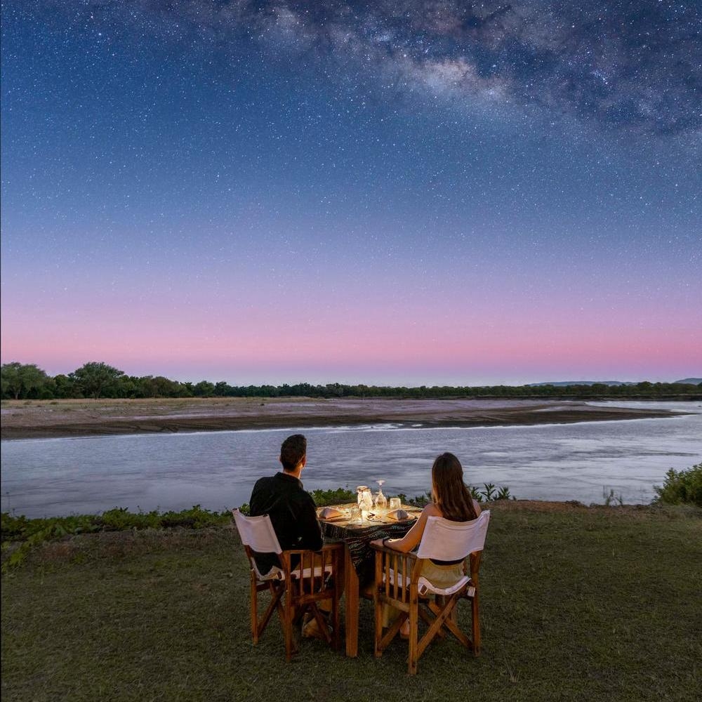 A couple sits at a dining table on a grassy riverbank under a clear night sky showing the Milky Way.
