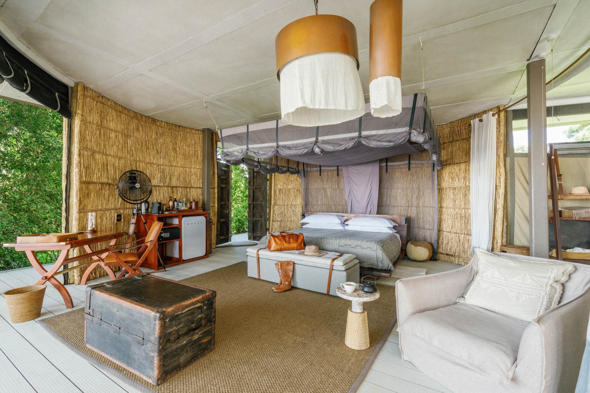 Interior of a luxury safari room with woven walls, a large bed, a vintage trunk, a desk, and a lounge chair.
