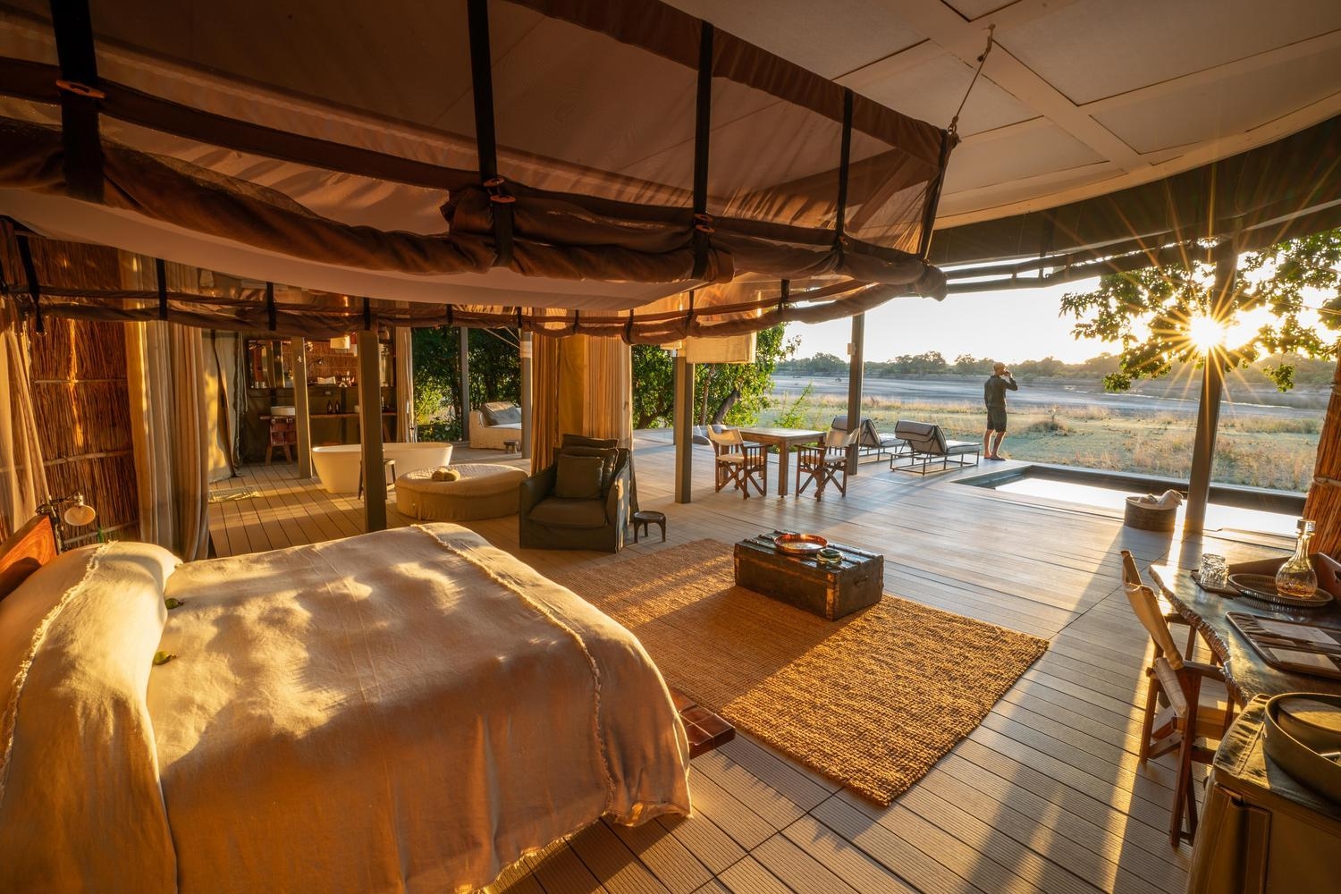 Interior of a luxury tent looking out to a deck where a man stands drinking, facing a river at sunrise.