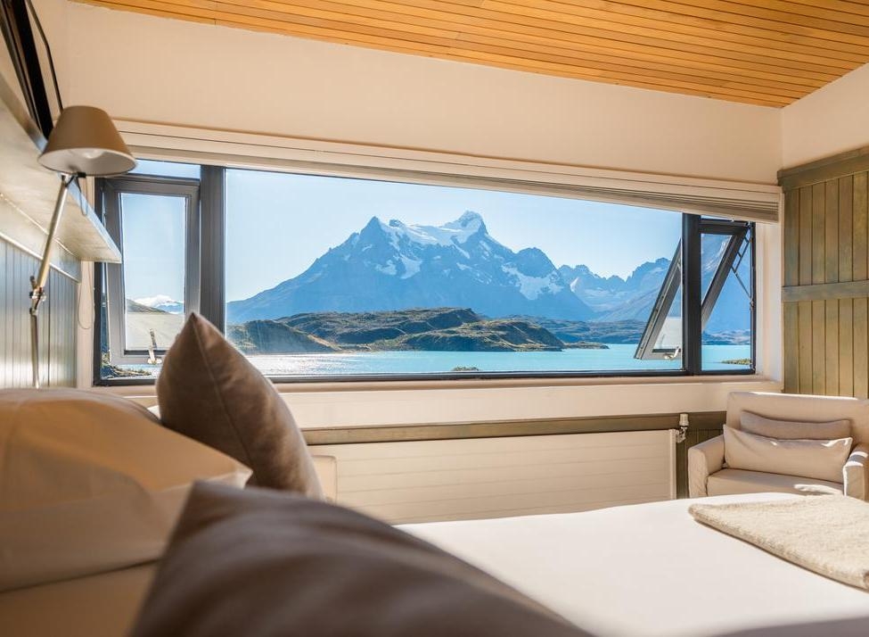 Large hotel window displaying a view of mountains and a bright blue lake from a bed in Explora Patagonia.