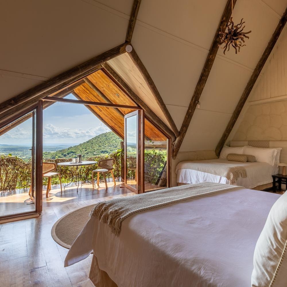 Bedroom with vaulted wood ceilings, two beds, and open glass doors leading to a private balcony overlooking a green landscape.