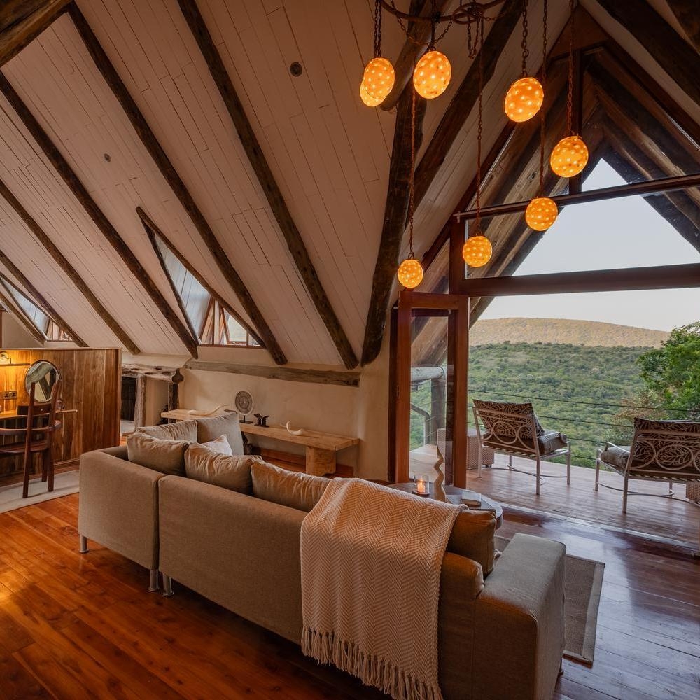Spacious bedroom lounge with vaulted ceilings, exposed beams, a large sofa, and an open doorway to a deck overlooking a valley.