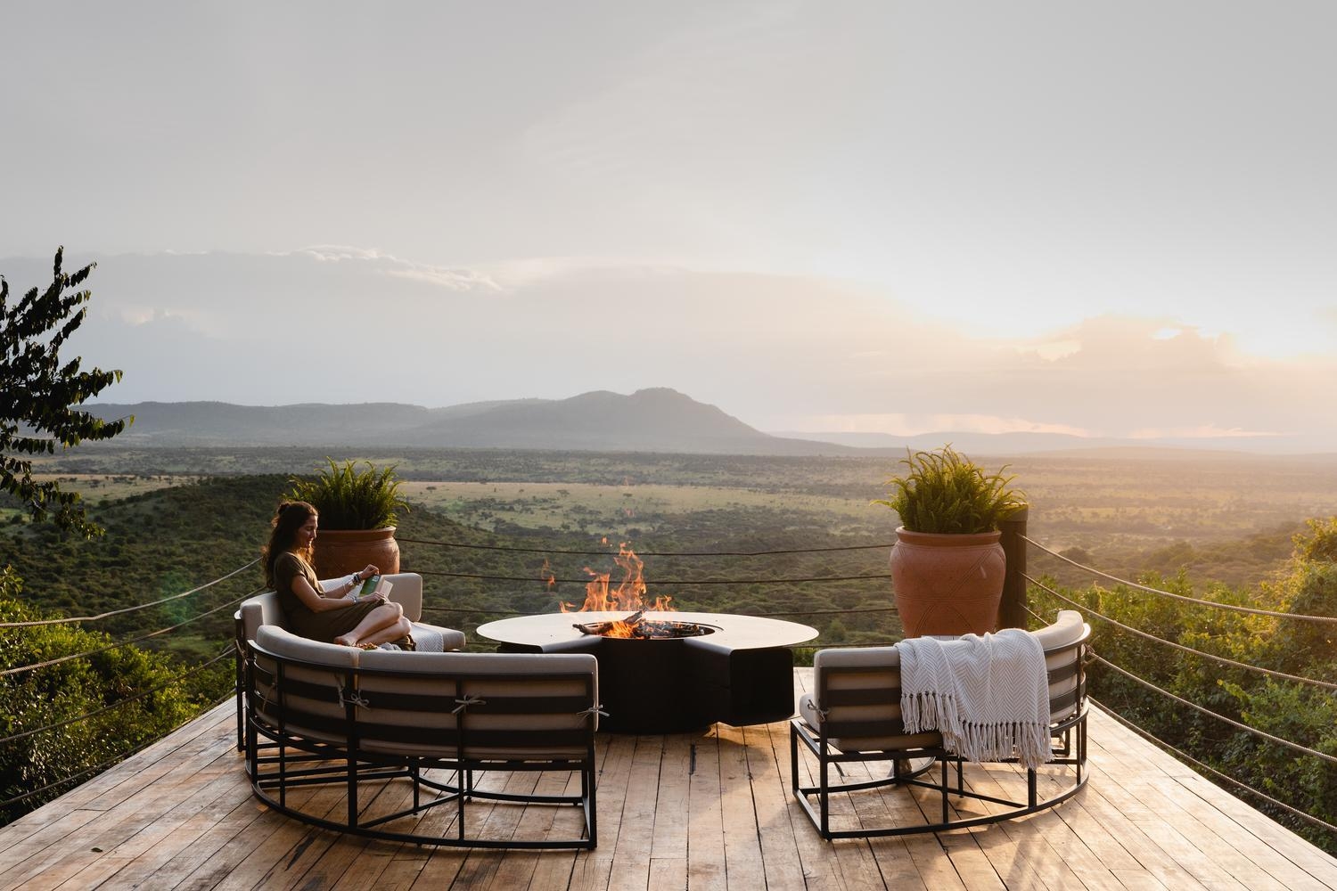 A woman reading on a curved outdoor sofa around a burning fire pit on a wooden deck, overlooking an expansive savanna.