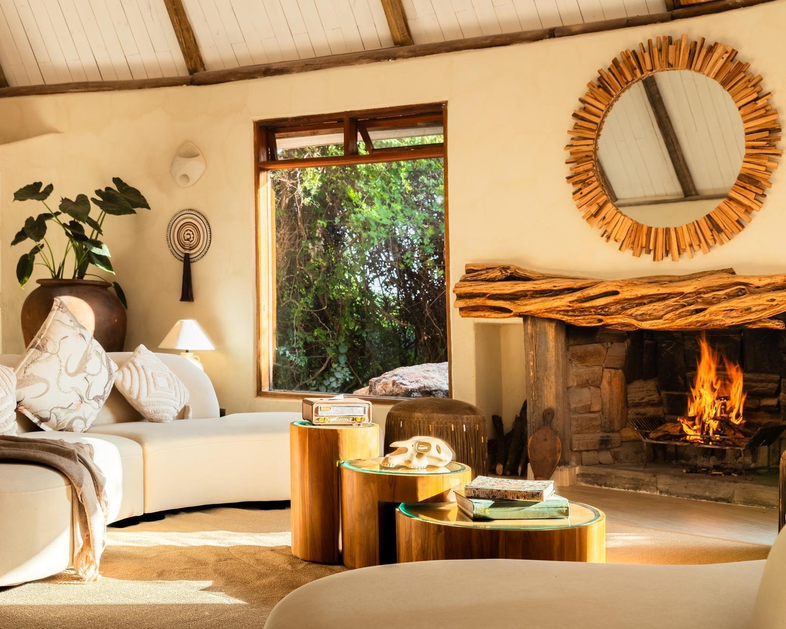 Cozy lounge with a curved white sofa, round wooden tables, and a burning fireplace featuring a rustic wood mantelpiece.
