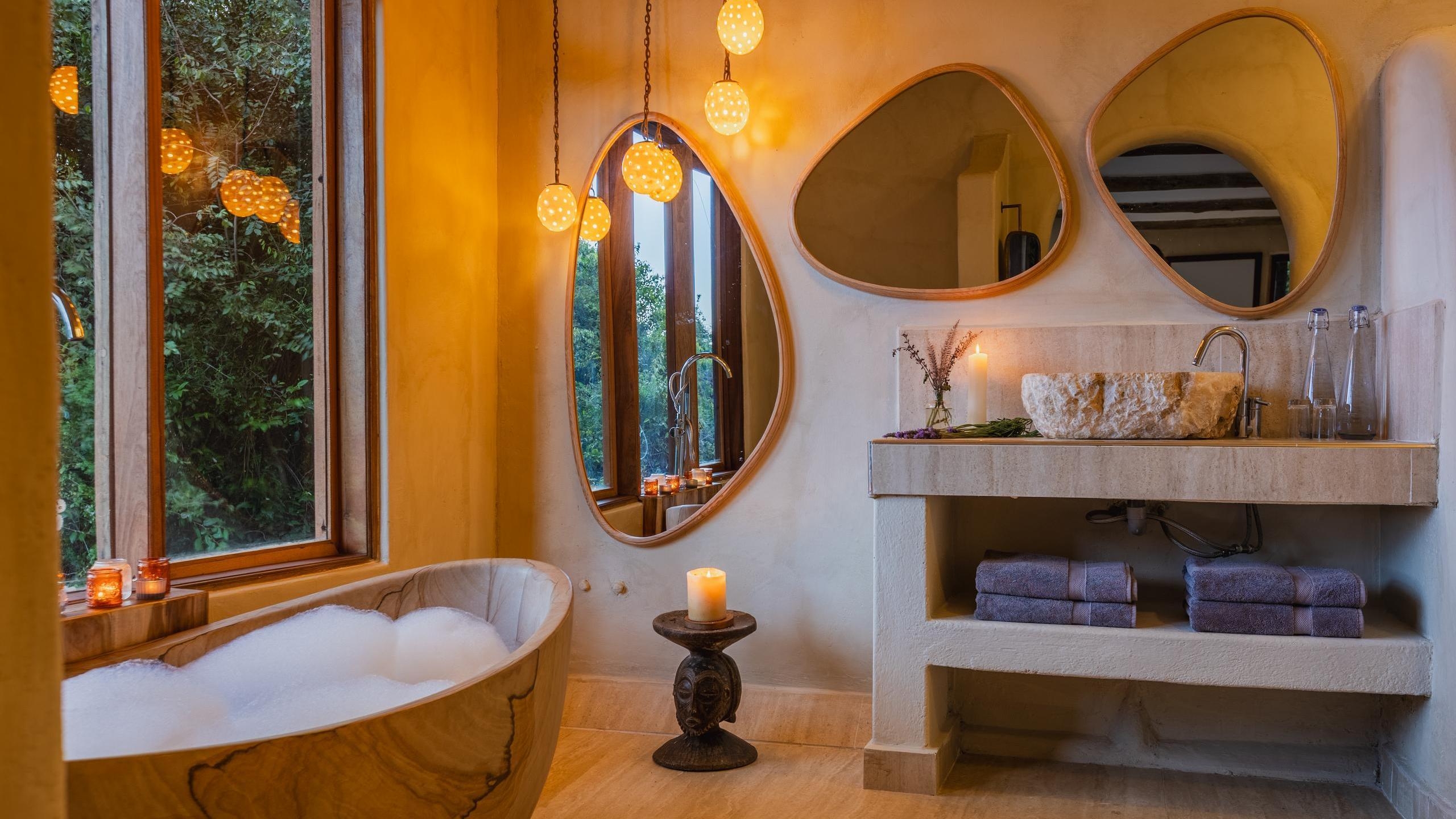 Rustic bathroom with a wooden bubble-filled soaking tub, stone sink, asymmetrical mirrors, and a window showing green foliage.