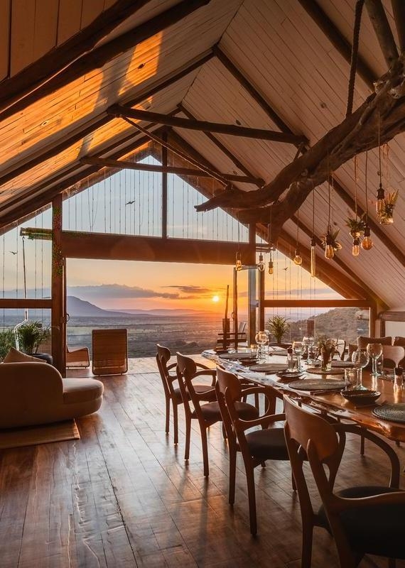 Dining room with a set wooden table, hanging lights, and large open windows framing a sunset over the savanna.