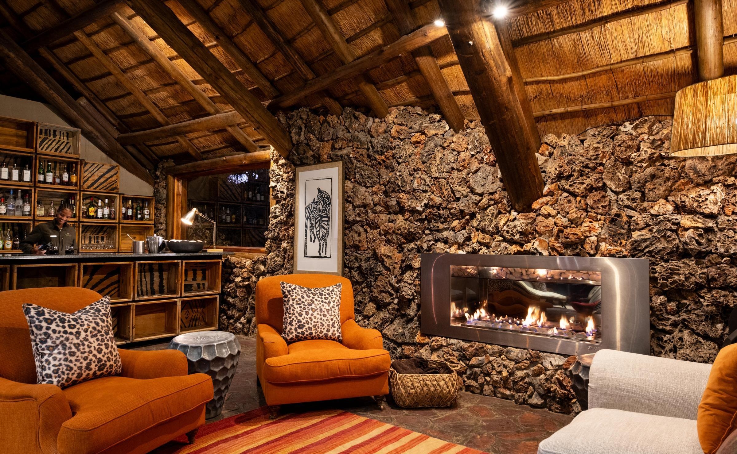 Cozy lounge at Ongava Lodge with a stone wall, modern fireplace, orange armchairs, and a thatched ceiling.