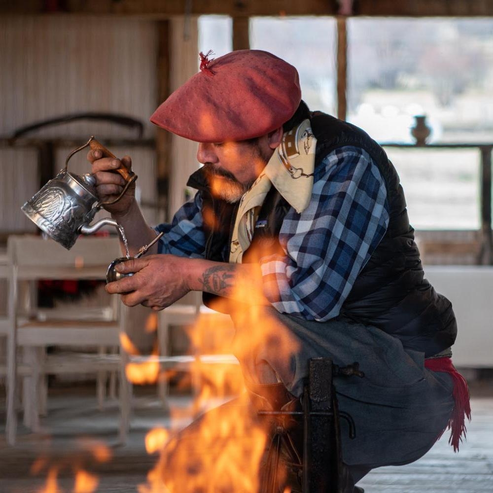 Man in a pink beret pouring water from an ornate silver kettle near a fire at Explora Patagonia.