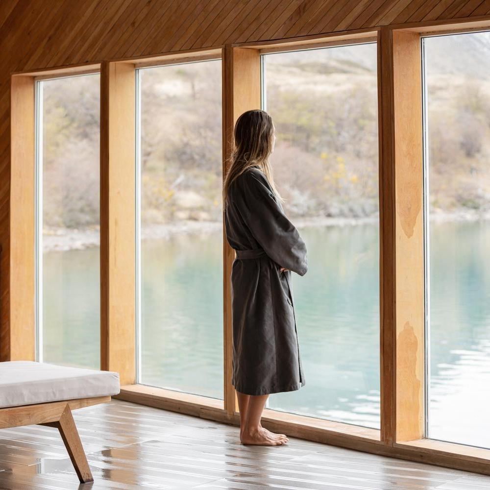 Person in a dark grey robe looking through large wooden-framed windows at a calm lake at Explora Patagonia.