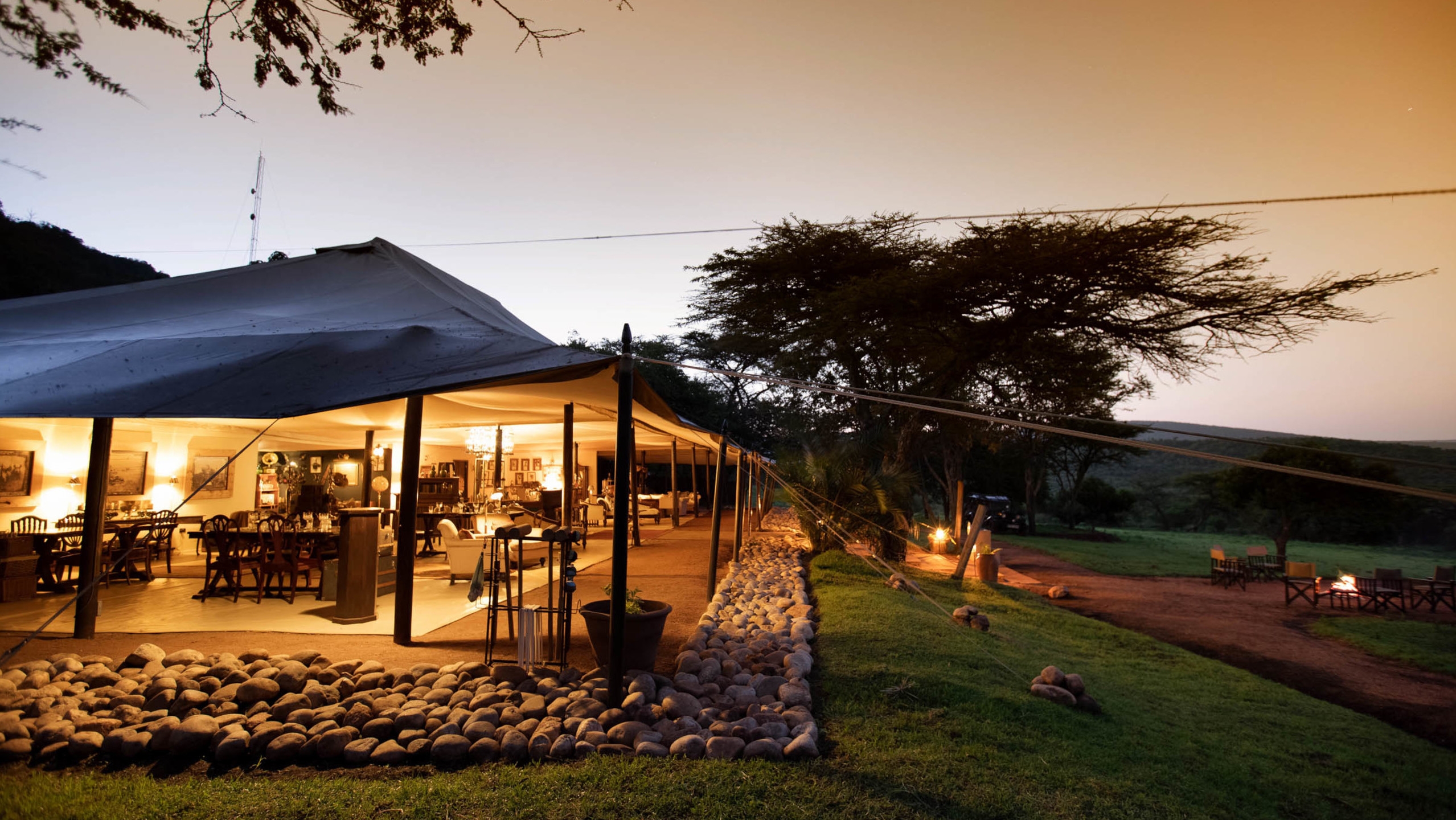 A large, brightly lit canvas safari mess tent at dusk showing dining and lounge areas, with a fire pit outside on the grass.