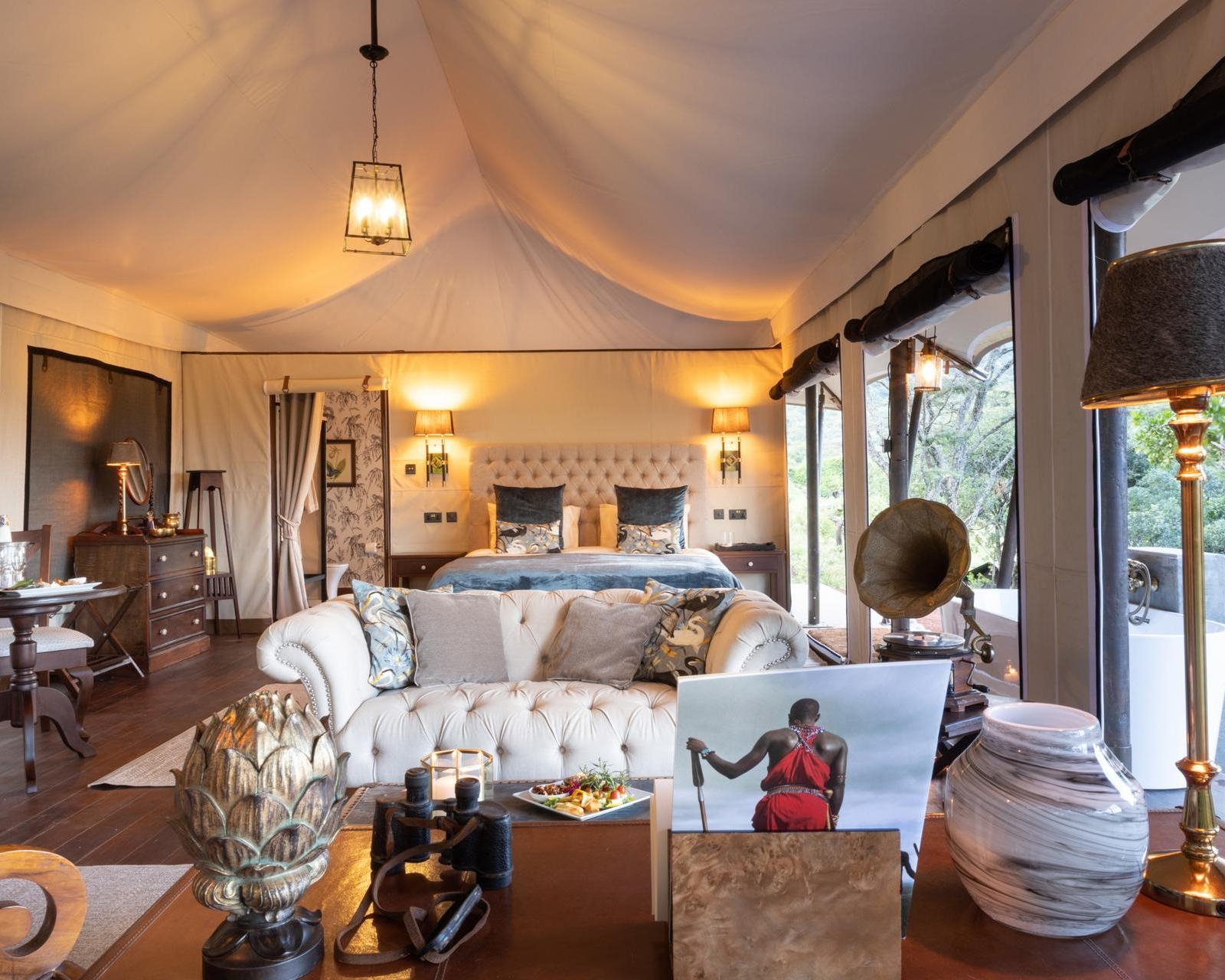 Lavish safari tent interior with a large bed, white tufted sofa, antique gramophone, and a book displaying a Maasai warrior.