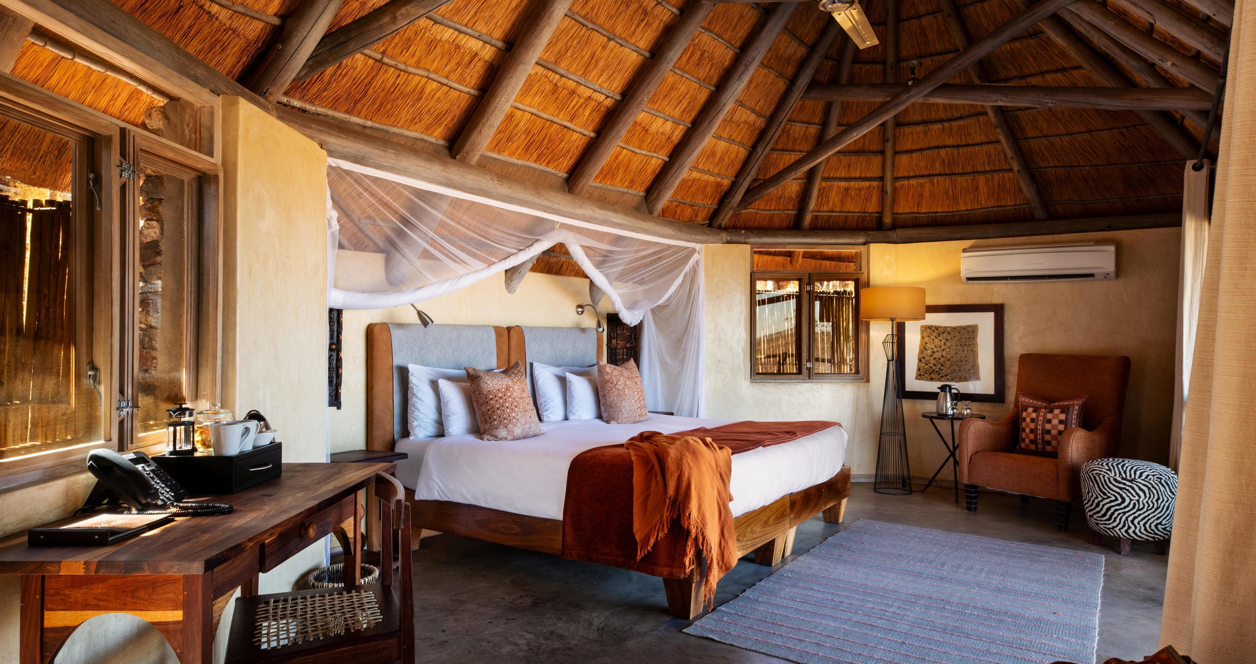 Luxury bedroom with a thatched roof, large bed with a mosquito net, and a wooden desk at Ongava Lodge.