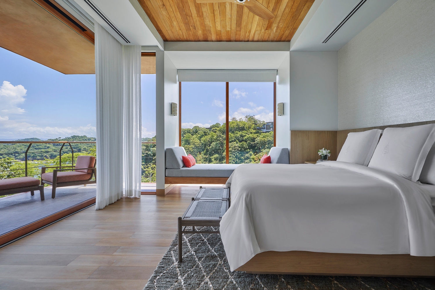 Bright modern bedroom with white linens, a wooden ceiling, and a large window showing a green forest view.