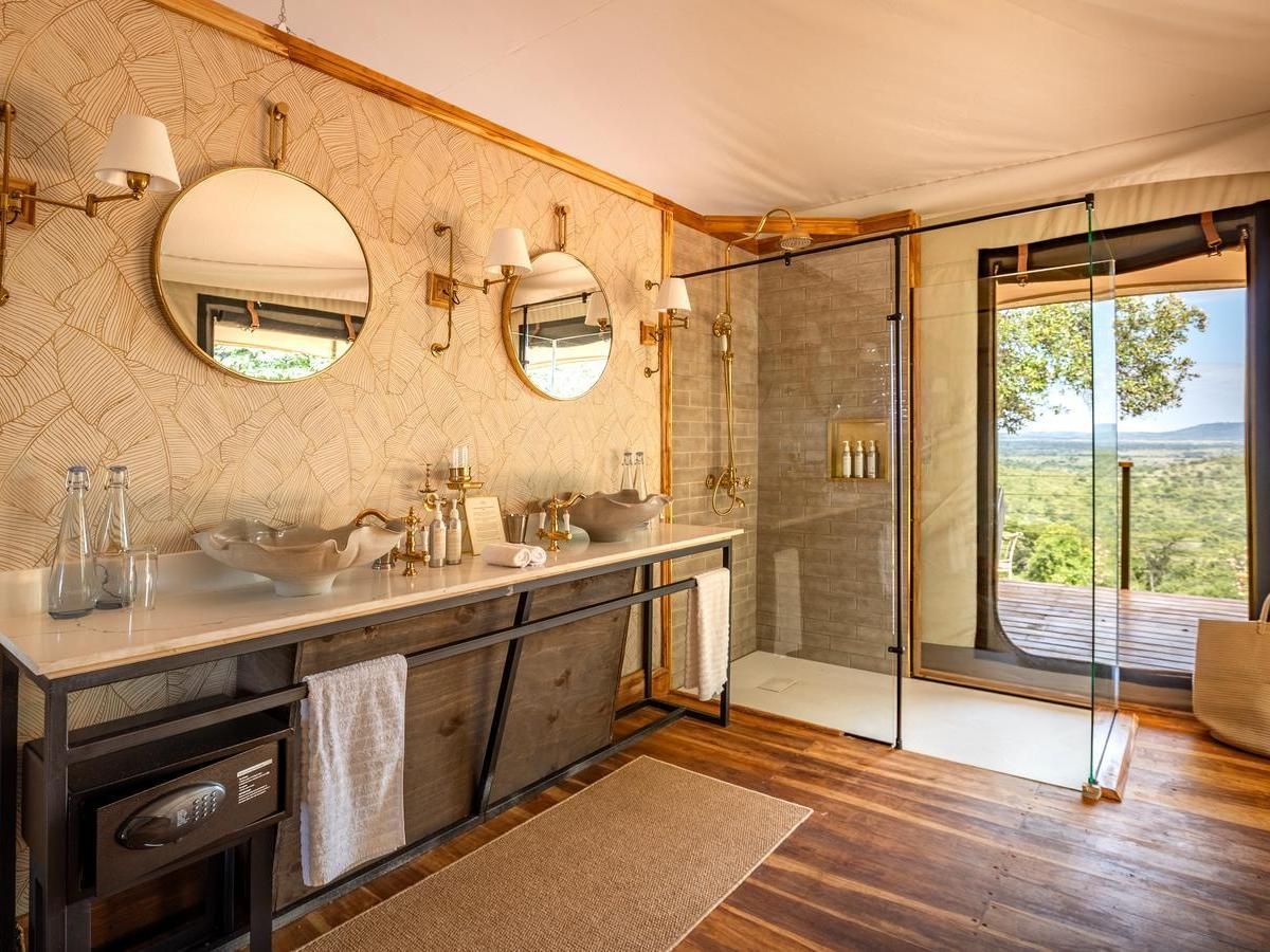 Luxury tent bathroom with double vanity, two round mirrors, leaf-patterned wallpaper, and a glass shower overlooking the savanna.