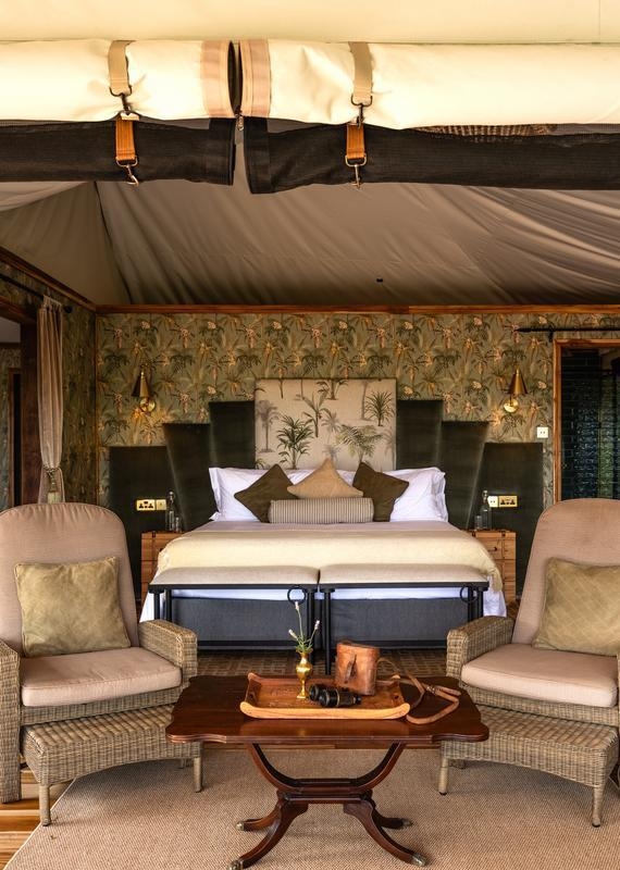 Safari tent bedroom with a large bed, tropical leaf patterned headboard, two wicker armchairs, and a coffee table with binoculars.