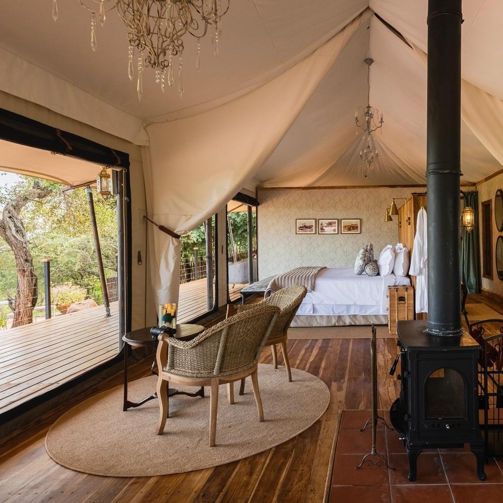 Luxury safari tent interior featuring a bed, two wicker chairs, a wood-burning stove, and a large opening to a wooden deck.