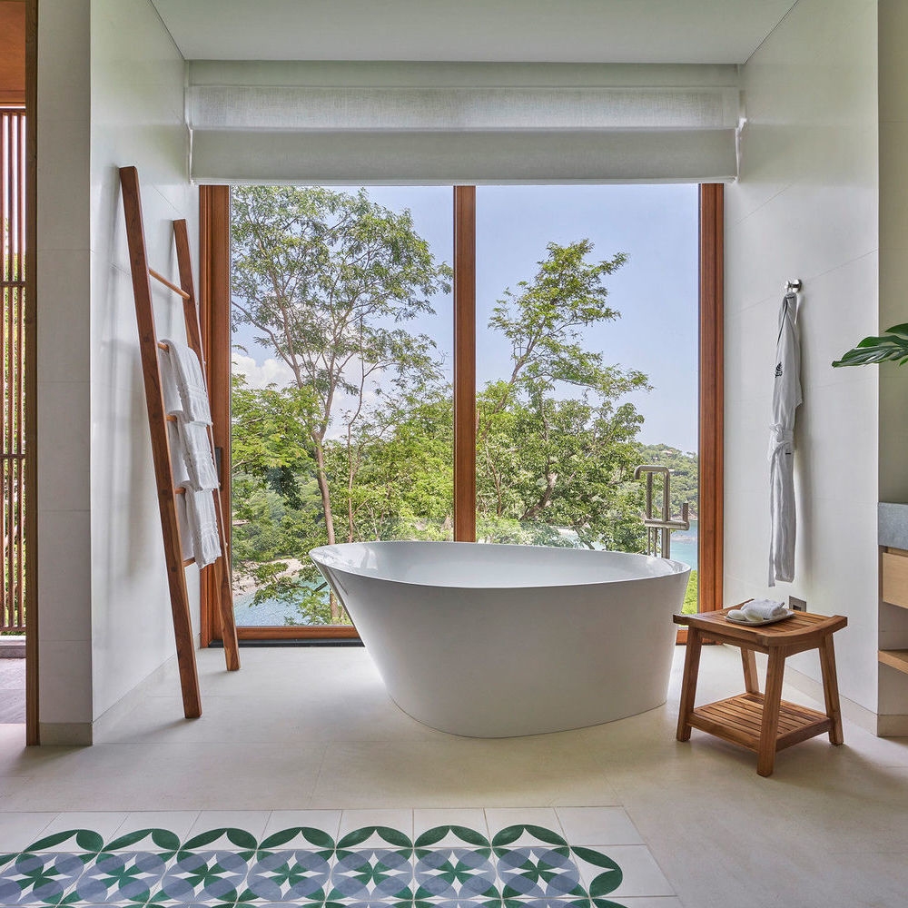 Modern white bathtub in front of a floor-to-ceiling window overlooking a tropical forest and the sea.